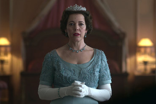 Elderly woman with brown hair and a blue dress wearing gloves and a crown and staring into the camera