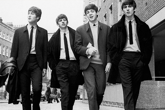 Four men with bowl cuts in suits walking towards the camera. Black and white photo