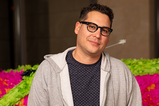 A man with glasses and gray jacket in front of piñata artwork