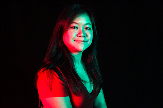 A woman with long black hair sitting against black background with colorful lighting