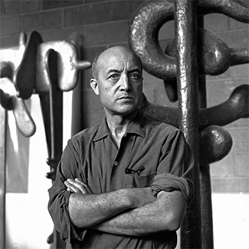 Black and white photo of artist Isamu Noguchi standing in front of his sculptures